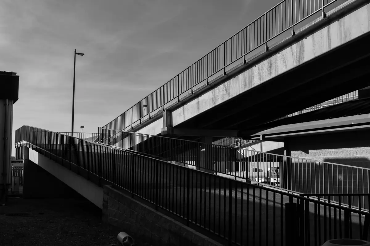 Passarela em rampa com guarda-corpo metálico sob um viaduto, em preto e branco