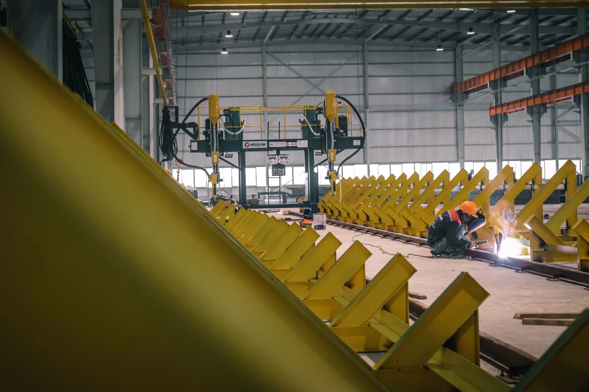 Soldador de capacete trabalhando junto a berços amarelos de apoio de vigas em galpão de fabricação, com pórtico de solda automática ao fundo