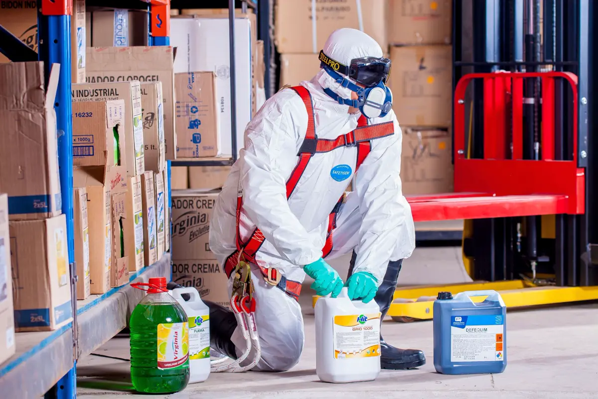 Trabalhador com macacão de proteção, óculos e luvas manuseando galões de produto químico num almoxarifado