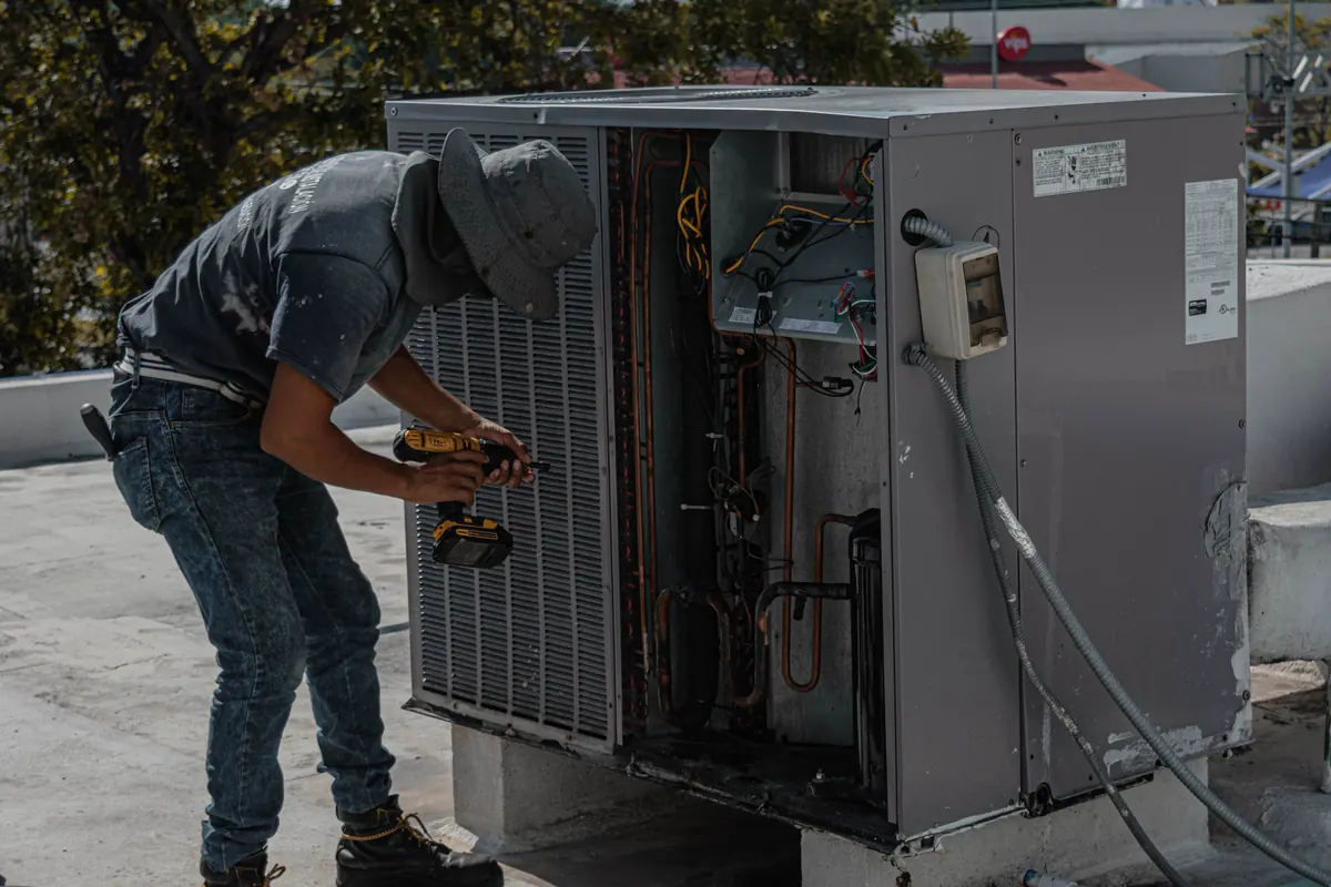 Técnico com parafusadeira trabalhando em unidade condensadora aberta, com tubulação de cobre e fiação do quadro expostas