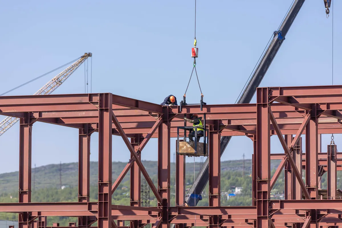 Montadores com capacete e colete trabalhando no alto de estrutura metálica em primer vermelho, com cesto de trabalho suspenso por guindaste e grua ao fundo