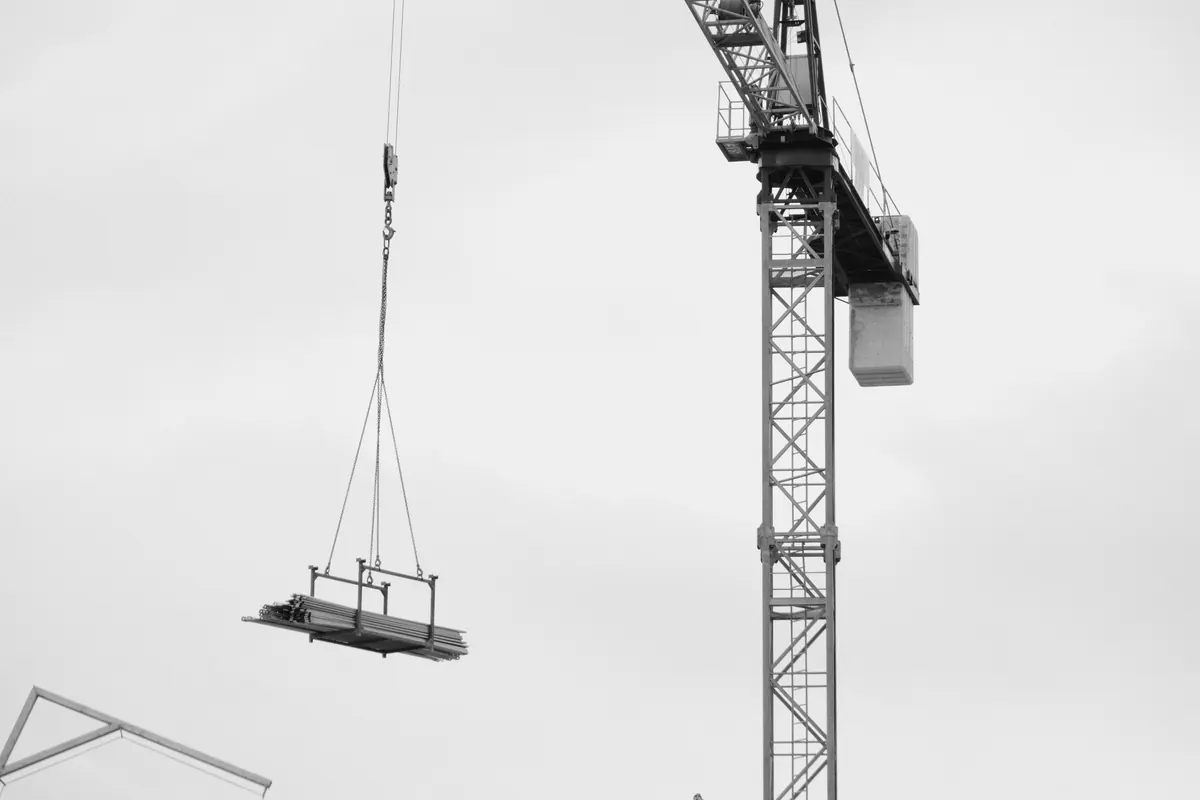 Grua de torre içando um fardo de perfis metálicos preso a um balancim, suspenso por lingas de corrente