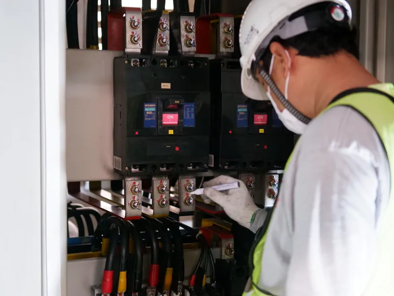 Técnico com capacete e colete refletivo inspecionando disjuntores e barramento dentro de painel elétrico industrial