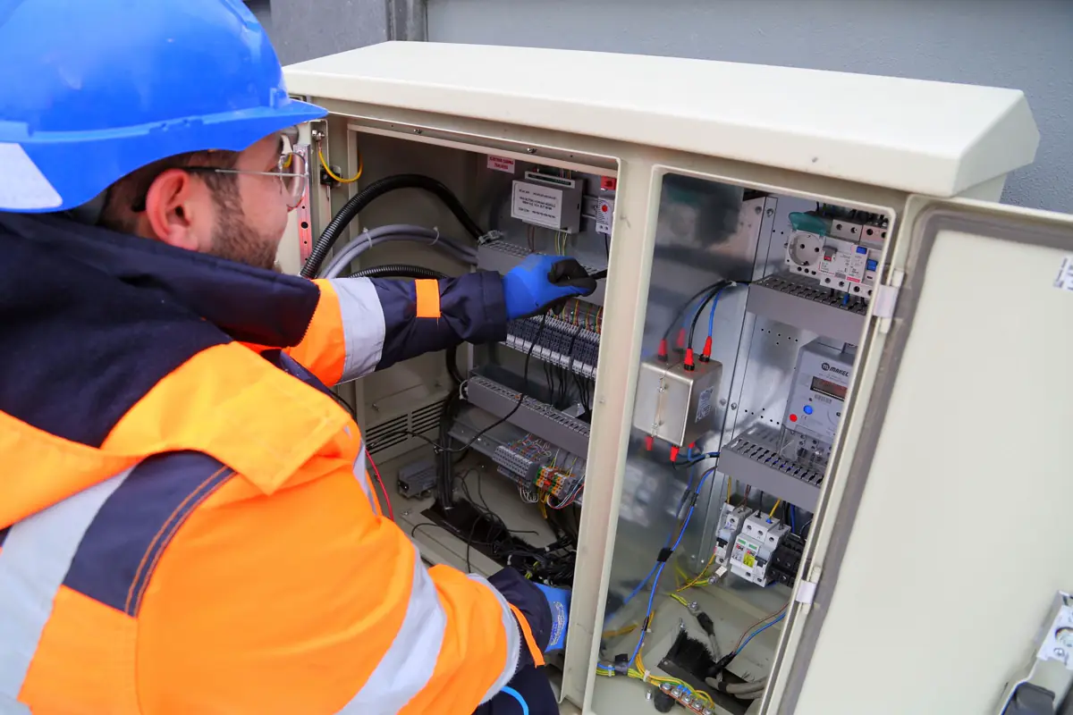 Eletricista de capacete e luvas mexendo nos disjuntores de um quadro elétrico externo