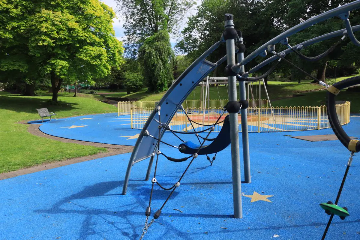 Brinquedo de escalar instalado sobre piso emborrachado azul em playground público, com balanços cercados ao fundo
