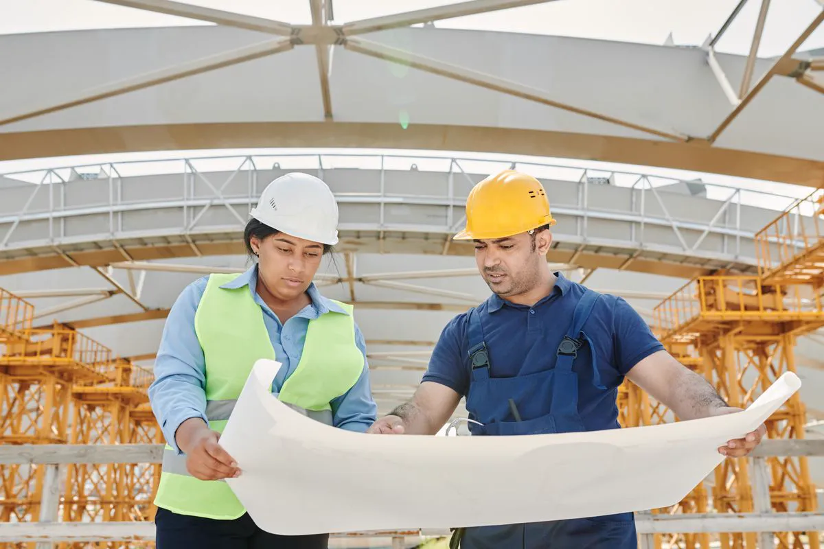 Engenheiros com capacete de segurança analisando projeto impresso de instalações em canteiro de obra