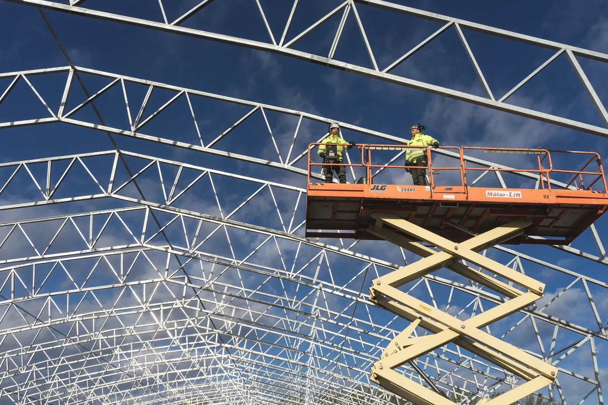 Dois montadores com capacete e colete em plataforma pantográfica durante a montagem de treliças metálicas em arco