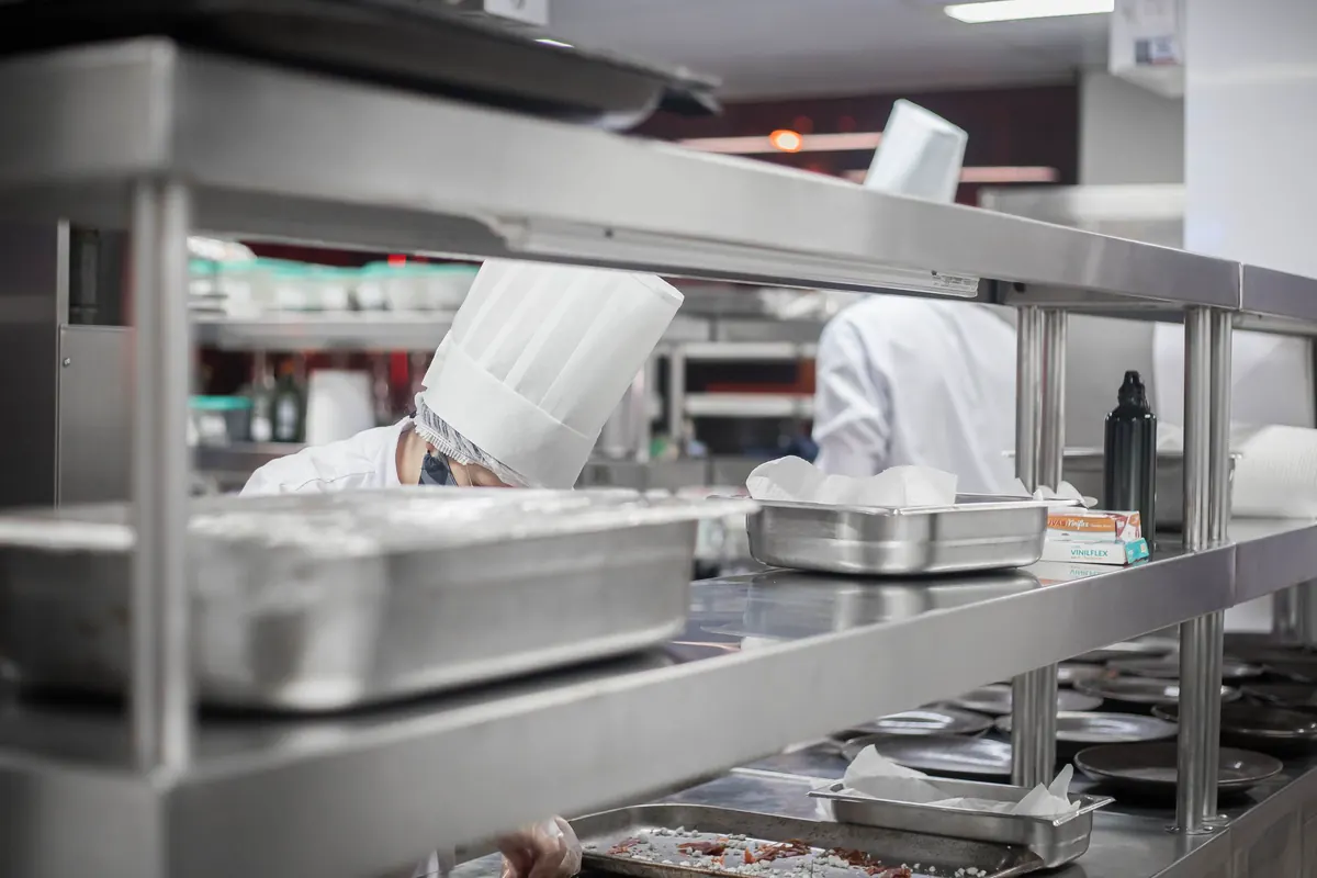 Cozinheiros de touca trabalhando atrás de prateleiras de inox com cubas gastronômicas em cozinha industrial