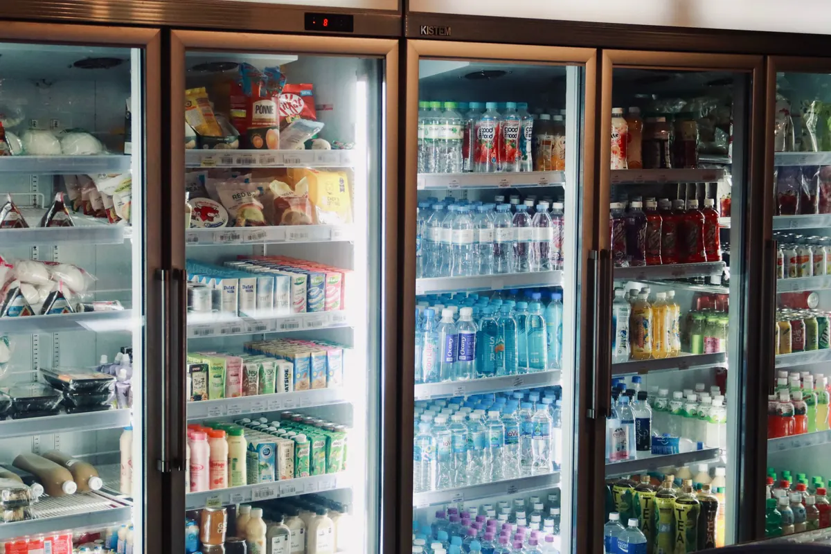 Fileira de refrigeradores de portas de vidro em loja, com bebidas, laticínios e alimentos embalados nas prateleiras